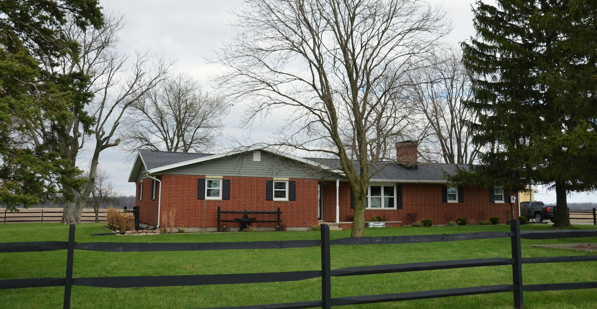 A red brick ranch-style house with mature trees and a black rail fence